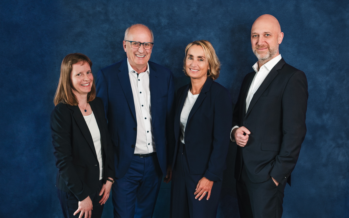 Vier Personen in Businesskleidung stehen nebeneinander vor einem blauen Hintergrund.